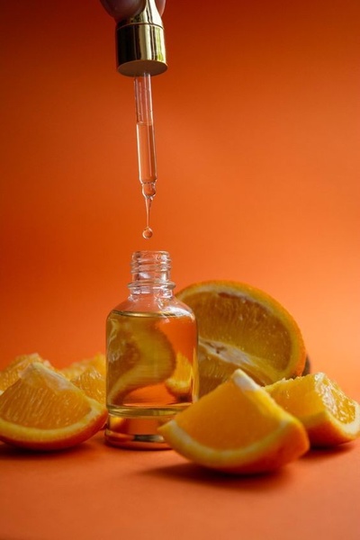 A clean, high-contrast product photography ad showing a serum dropper releasing a droplet above an open bottle surrounded by orange slices.